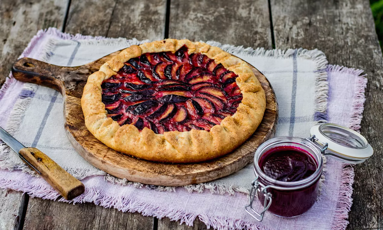 Галет със златисто-кафява коричка, подредени резени от тъмно виолетови сливи и преливен слой червено сладко върху дървена плоча