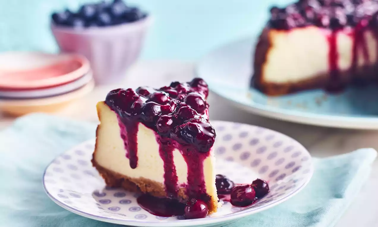 Cheesecake s hladkým světlým vanilkovým krémem na zlatavé sušenkové základně, přelitý tmavě fialovou borůvkovou omáčkou na puntíkovaném talíři