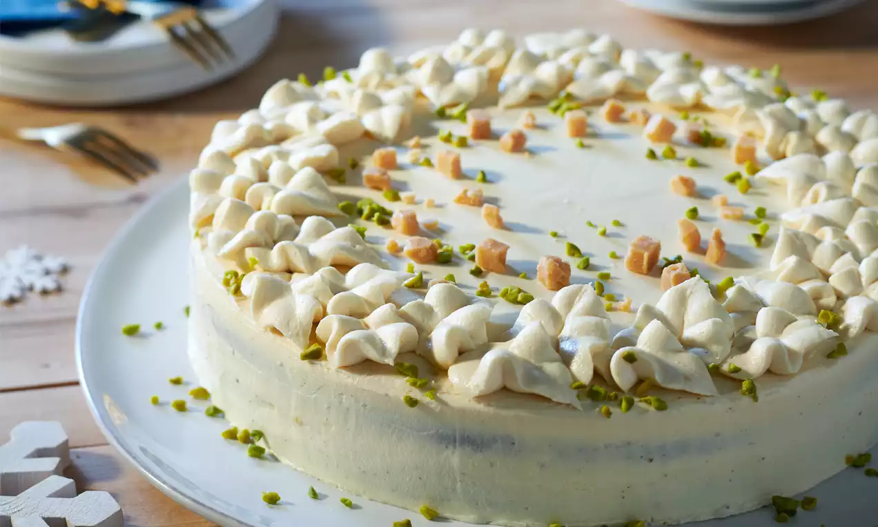 Krémfehér torta vaníliás bevonattal, tejszínhabos csúcsokkal, karamellkockákkal és zöld pisztáciadarabokkal díszítve fehér tálon, faasztalon