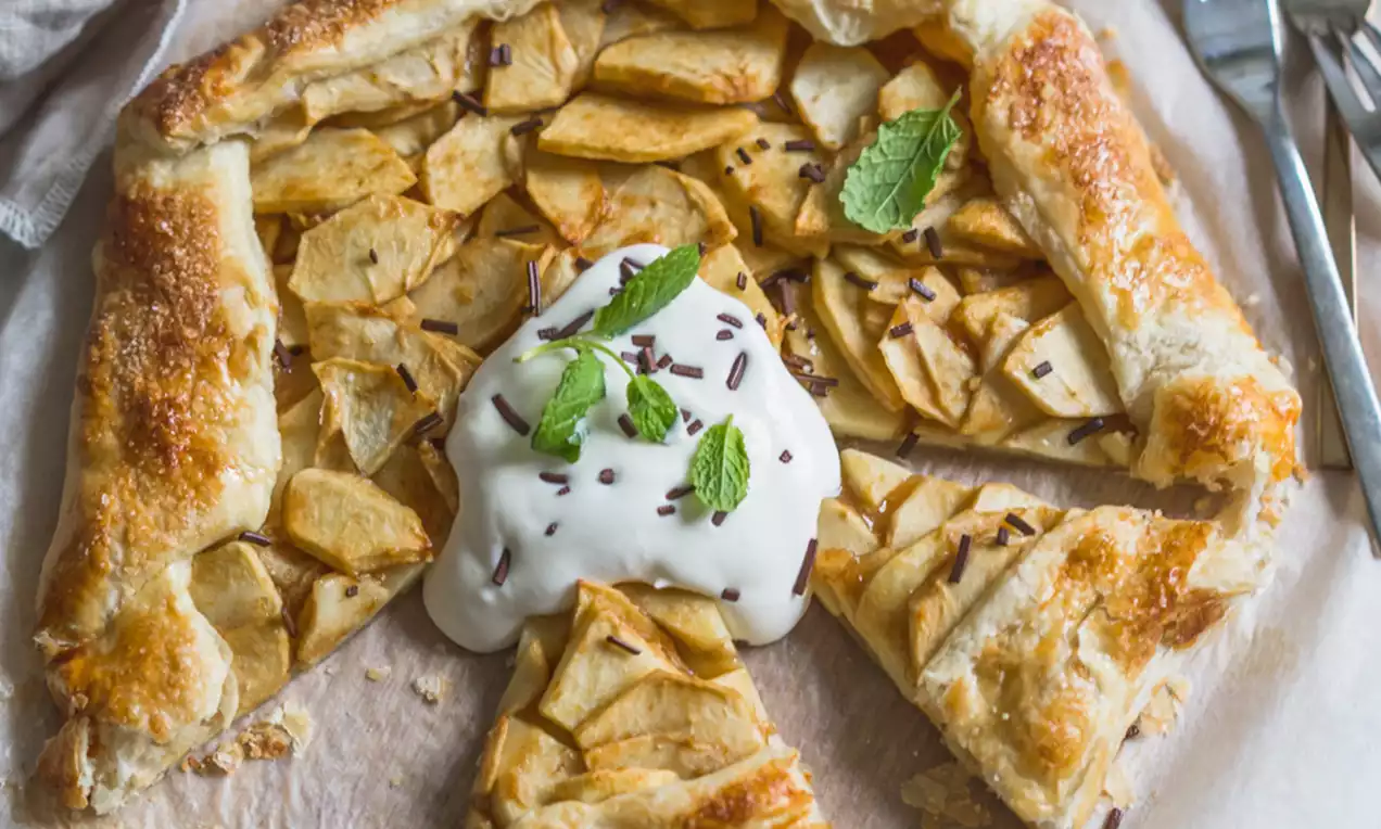 Tarte rustique aux pommes dorées, bordure croustillante, garnie de crème blanche, copeaux de chocolat et feuilles de menthe sur papier cuisson