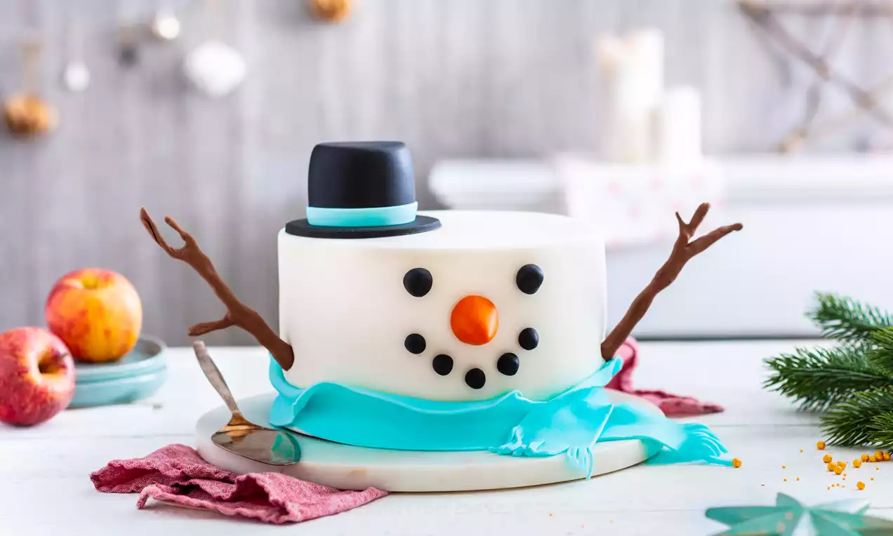 Gâteau en forme de bonhomme de neige blanc avec un chapeau noir, branches en guise de bras, écharpe bleue et détails en pâte colorée sur une assiette