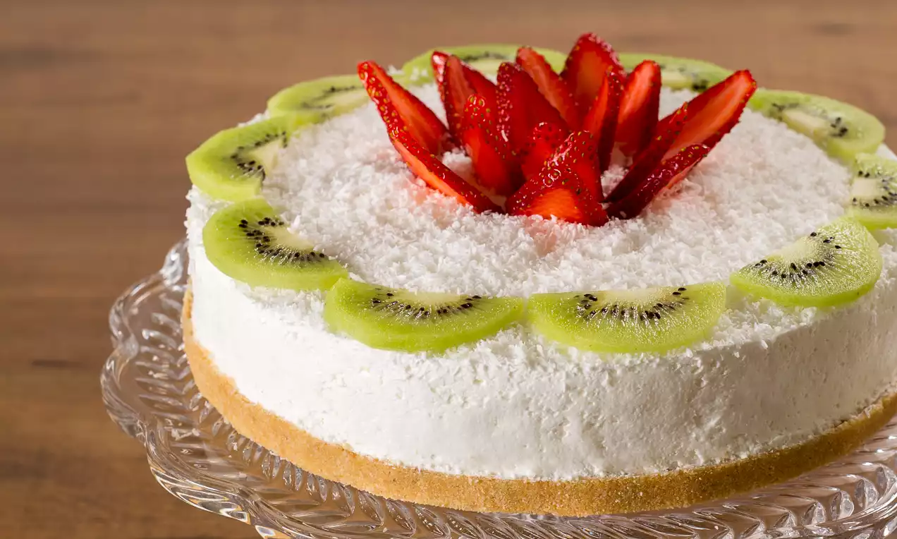 Torta con base di biscotto dorata, crema bianca soffice ricoperta di cocco e decorata con fettine di kiwi e fragole disposte a fiore