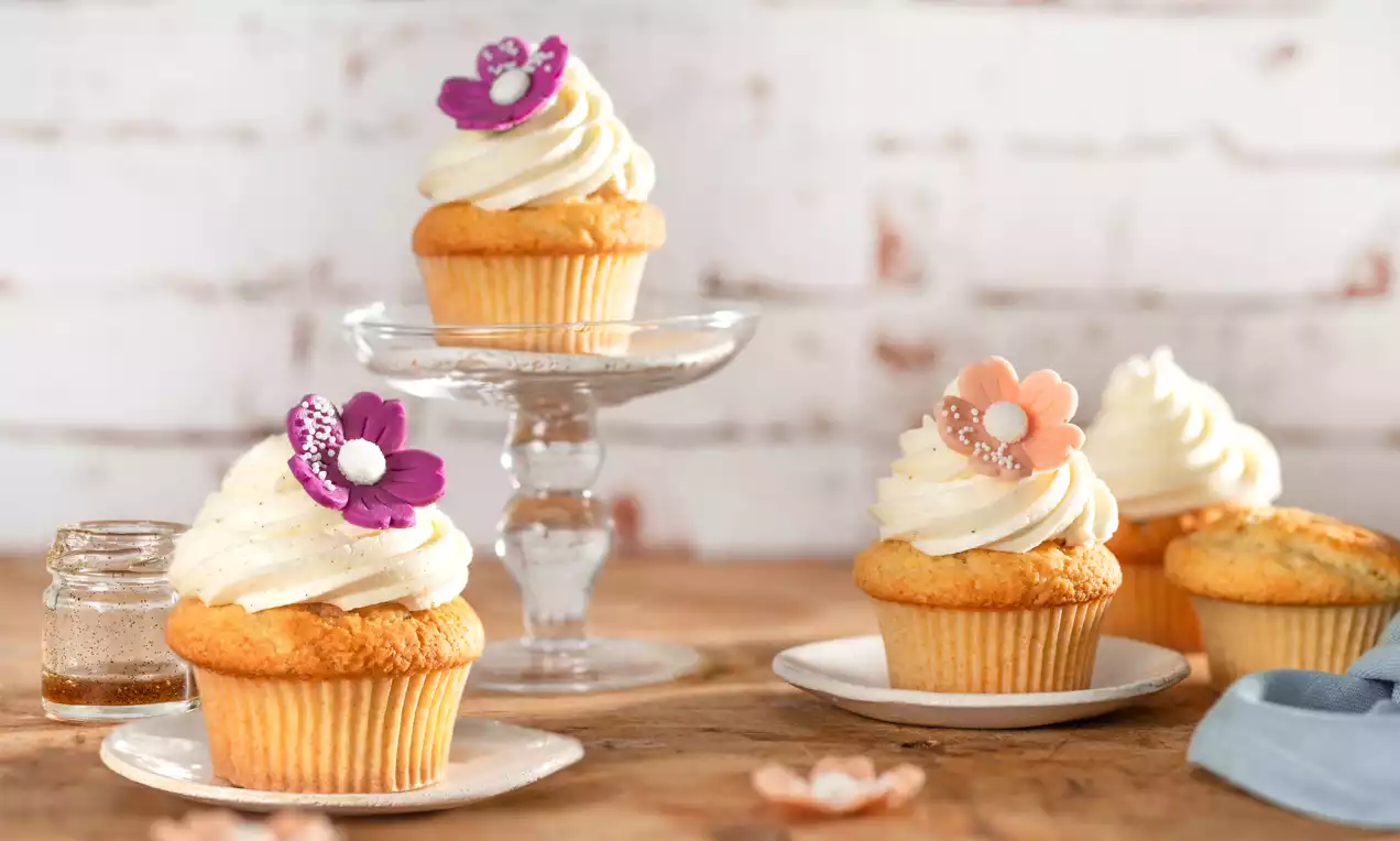 Goldgelbe Vanille-Cupcakes mit geschwungener weißer Cremehaube und filigranen Blüten, arrangiert auf Tellern und einer Glas-Etagere