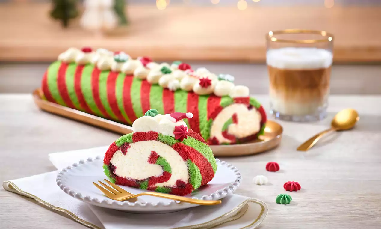 Božična biskvitna rulada z rdeče-zelenimi črtami, kremnim polnilom, okrašena z belimi in rdečimi kapicami na krožniku zlatih robov