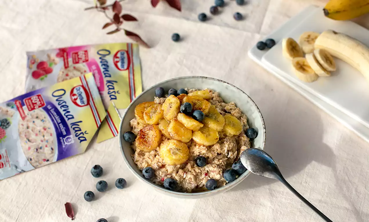 Bļodā ar krēmīgām, gaiši bēšām auzu pārslām kārtojas zeltaini karamelizēti banānu šķēlītes un sulīgas, tumši zilas mellenes
