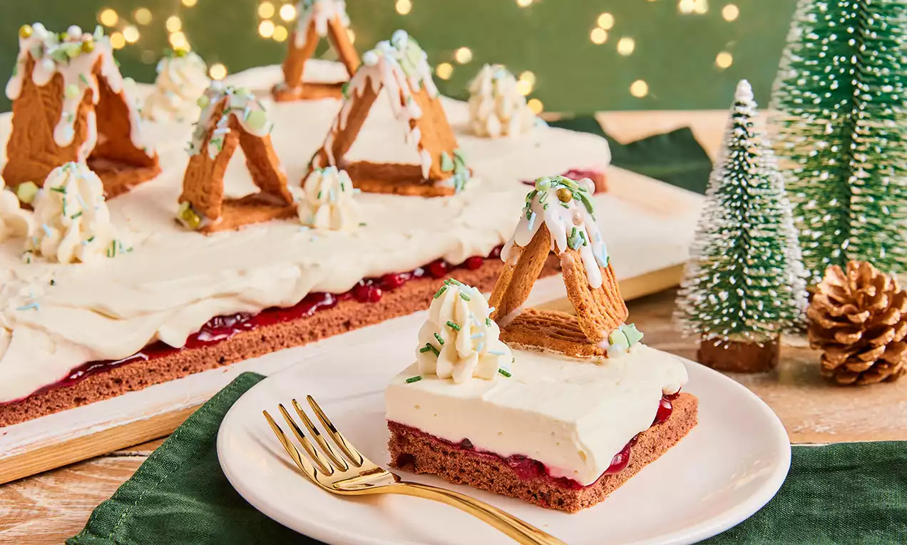 Rechteckiger Kuchen mit braunem Boden, weißer Creme, roter Schicht, Spekulatius-Häusern und Sahnetupfen, dekoriert mit Zuckerguss und Pistazien