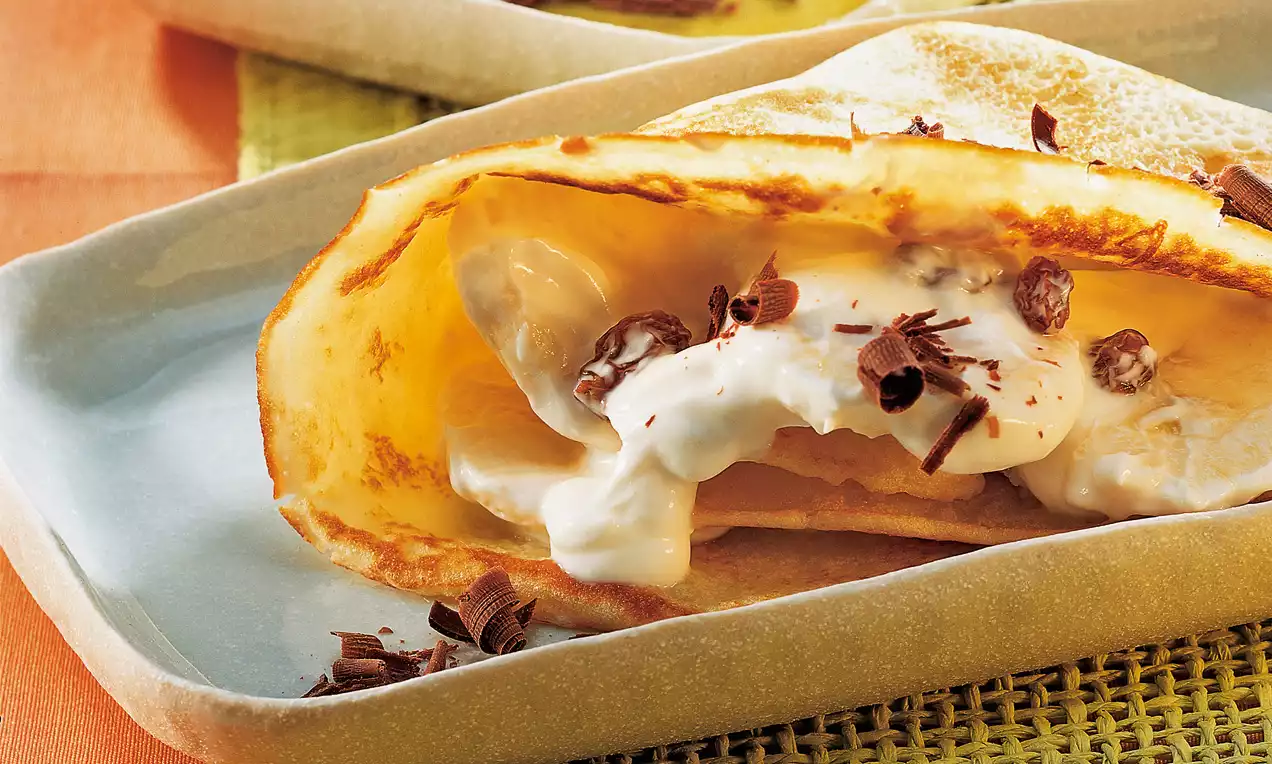 Crêpes dorées pliées garnies de crème blanche, morceaux de banane et copeaux de chocolat sur une assiette beige