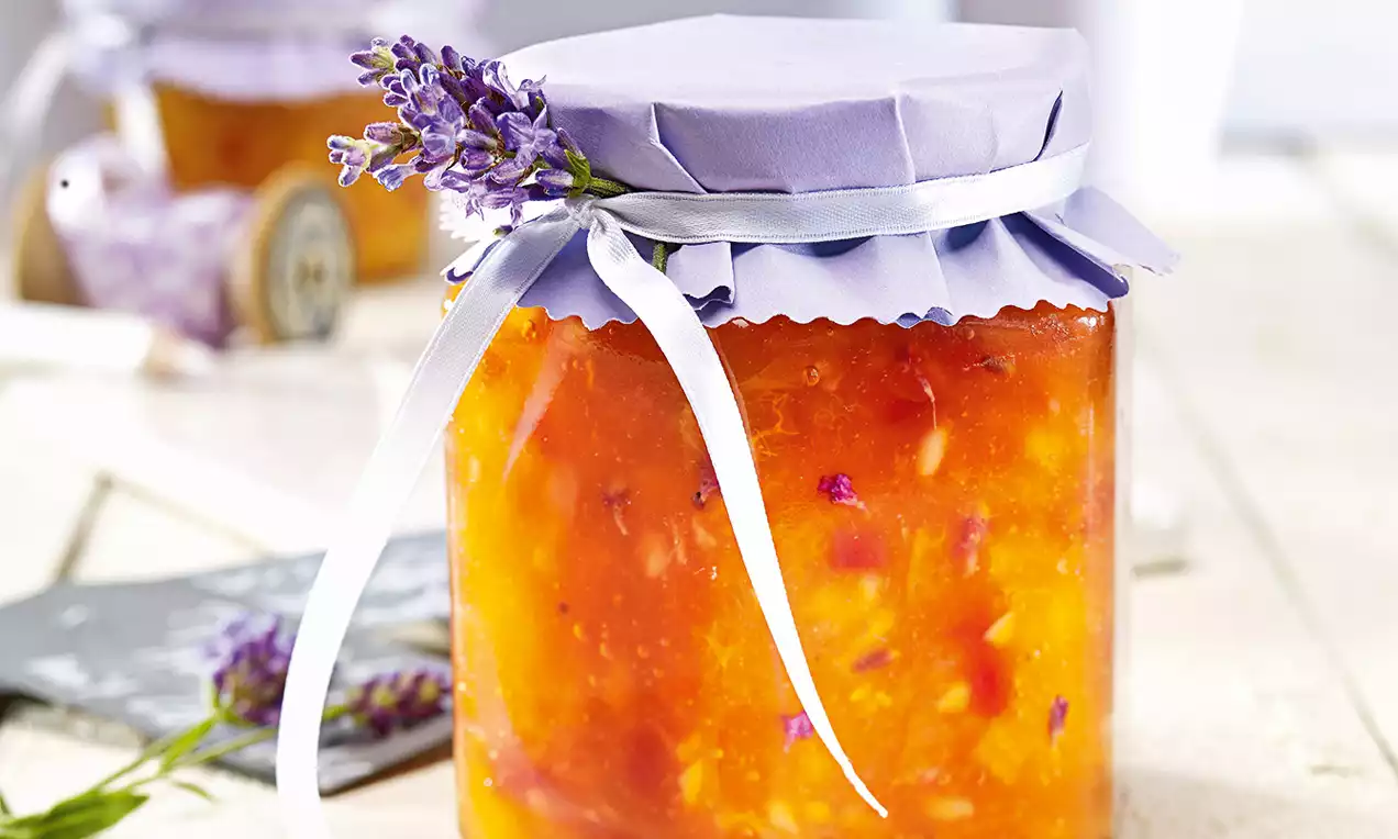 Confiture d'abricots orange ambrée parsemée de morceaux, dans un pot décoré d’un couvercle lavande noué d’un ruban blanc et brindille de lavande