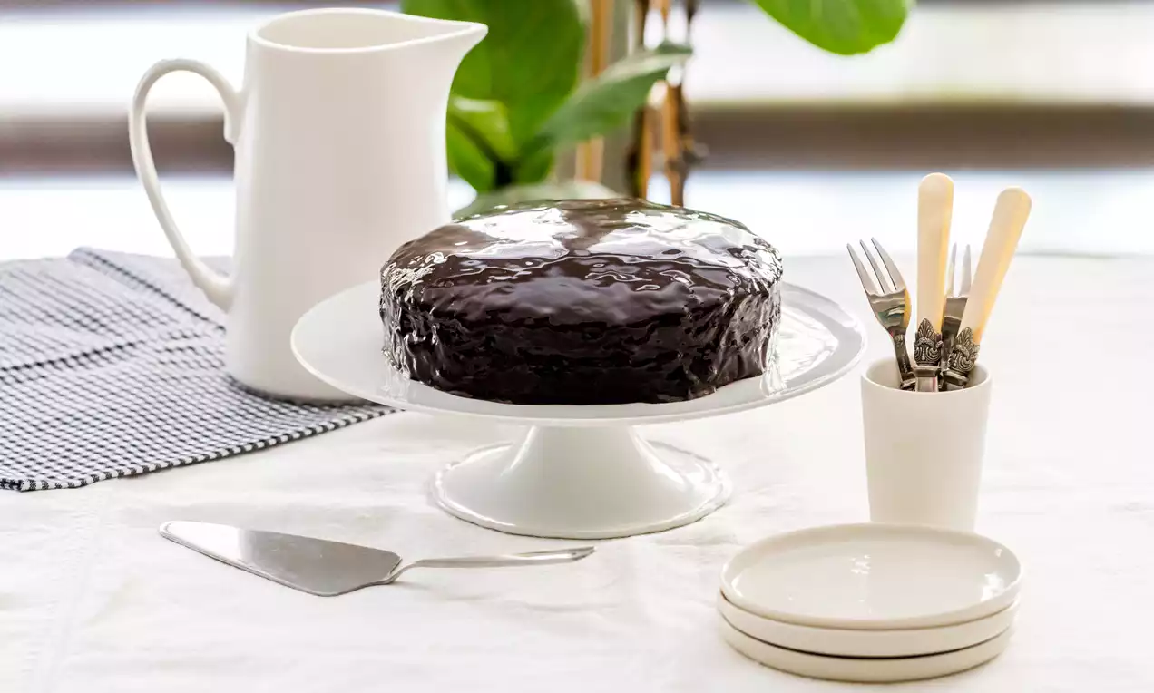 Tarta Sacher redonda cubierta de un glaseado brillante de chocolate oscuro sobre un plato blanco con cubiertos y vajilla clara alrededor