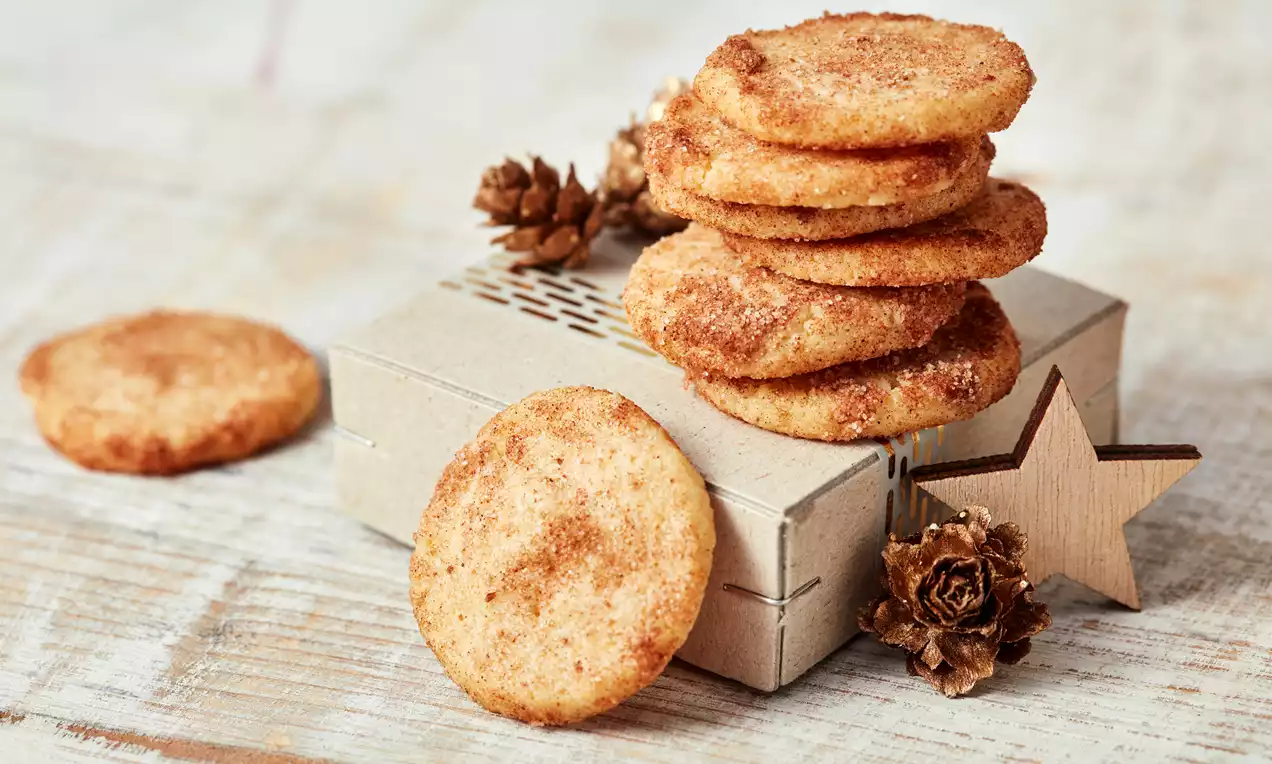 Goldbraune, runde Zimtkekse mit knusprigem Rand, teils gestapelt auf cremefarbener Schachtel mit Holzsternen und Deko-Tannenzapfen
