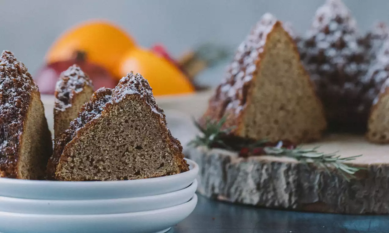 Porciones triangulares de pastel especiado marrón cubiertas con azúcar glas, dispuestas sobre un plato blanco junto a ramas de romero y rodajas de naranja