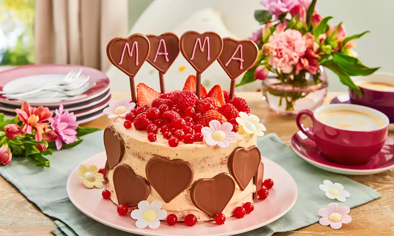 Cremefarbene Torte mit Schokoladenherzen, roten Beeren und Schokoladen-Aufstellern auf pastellrosafarbenem Teller mit Blumen dekoriert