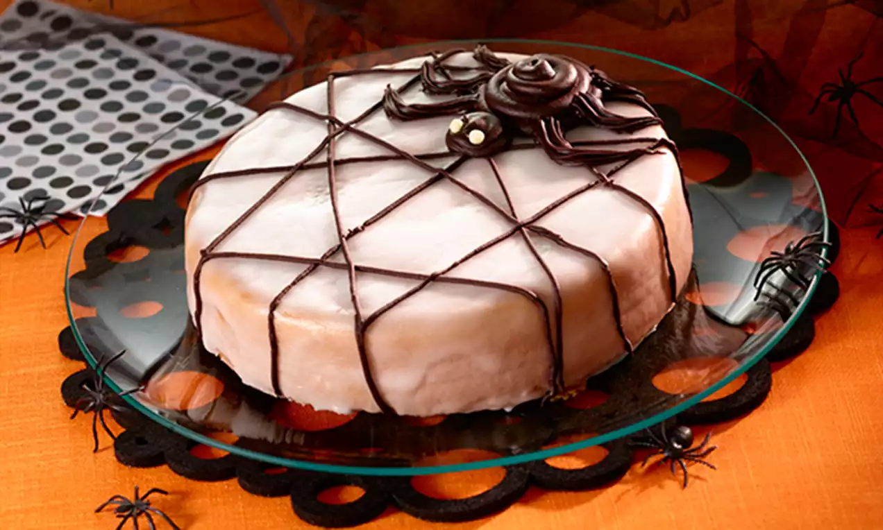 Gâteau rond glacé blanc surmonté d'une toile en chocolat noir avec une araignée sculptée posée au centre sur un plateau transparent