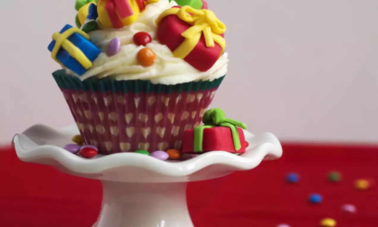 Sinterklaas-cupcake in rood-wit vormpje met romige witte topping, kleurrijke marsepeinen cadeautjes en suikerparels op een wit taartplateau