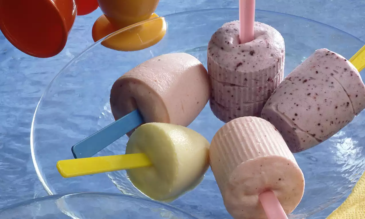 Farbig gemischte Eislutscher in runder, zylindrischer Form auf transparentem Glasteller mit sichtbaren Holz- und Plastestielen