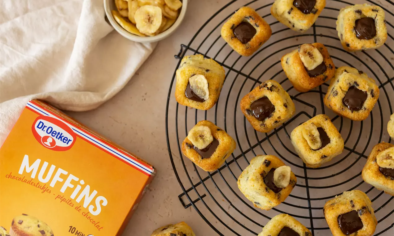 Petits muffins dorés à la banane et pépites de chocolat sur une grille noire avec rondelles de banane et carré de chocolat fondant