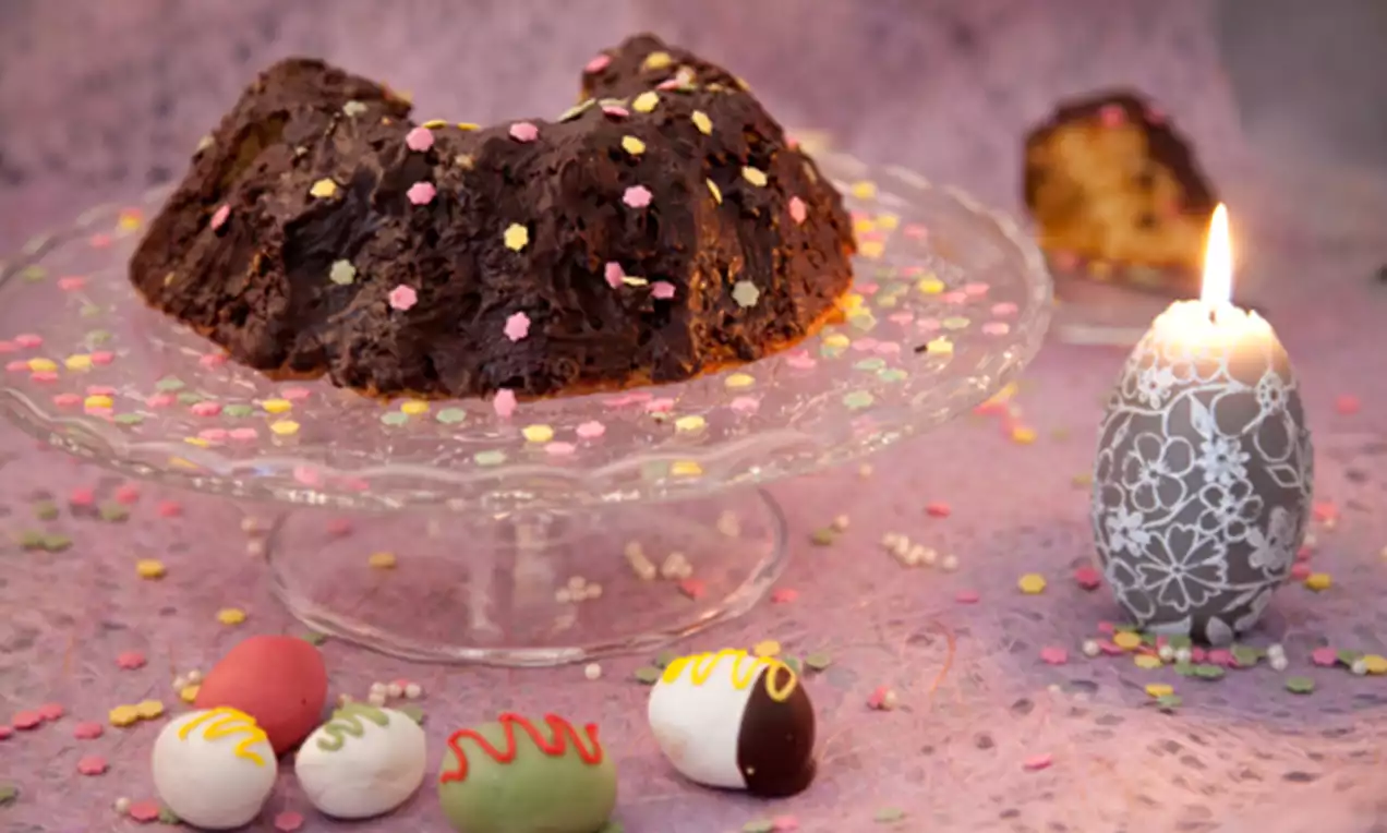 Fruktig påsktårta med mörk chokladglasyr och färgglada strössel på glasfat, omgiven av dekorerade ägg och ett tänd mönstrat ljus