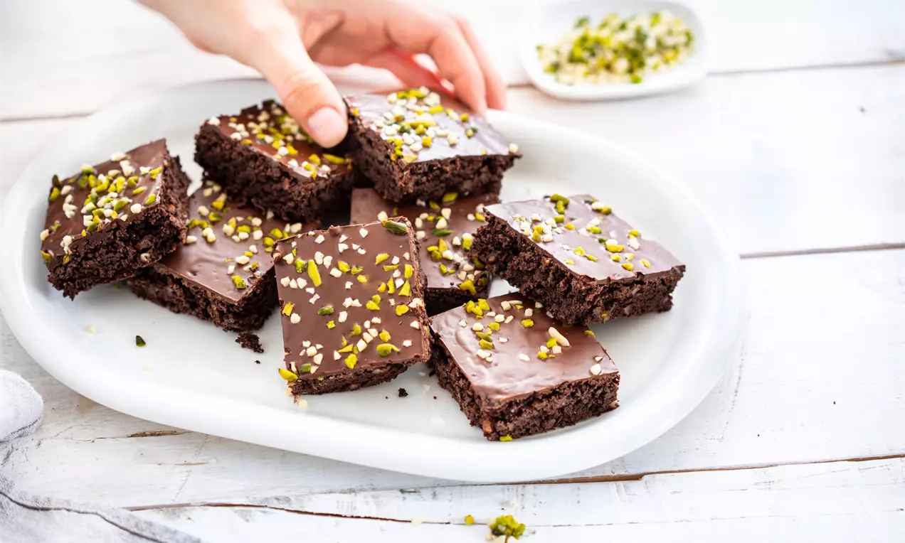Тъмнокафяви правоъгълни парчета брауни с гладка шоколадова глазура, поръсени с ситно нарязани шамфъстъци върху бяла овална чиния