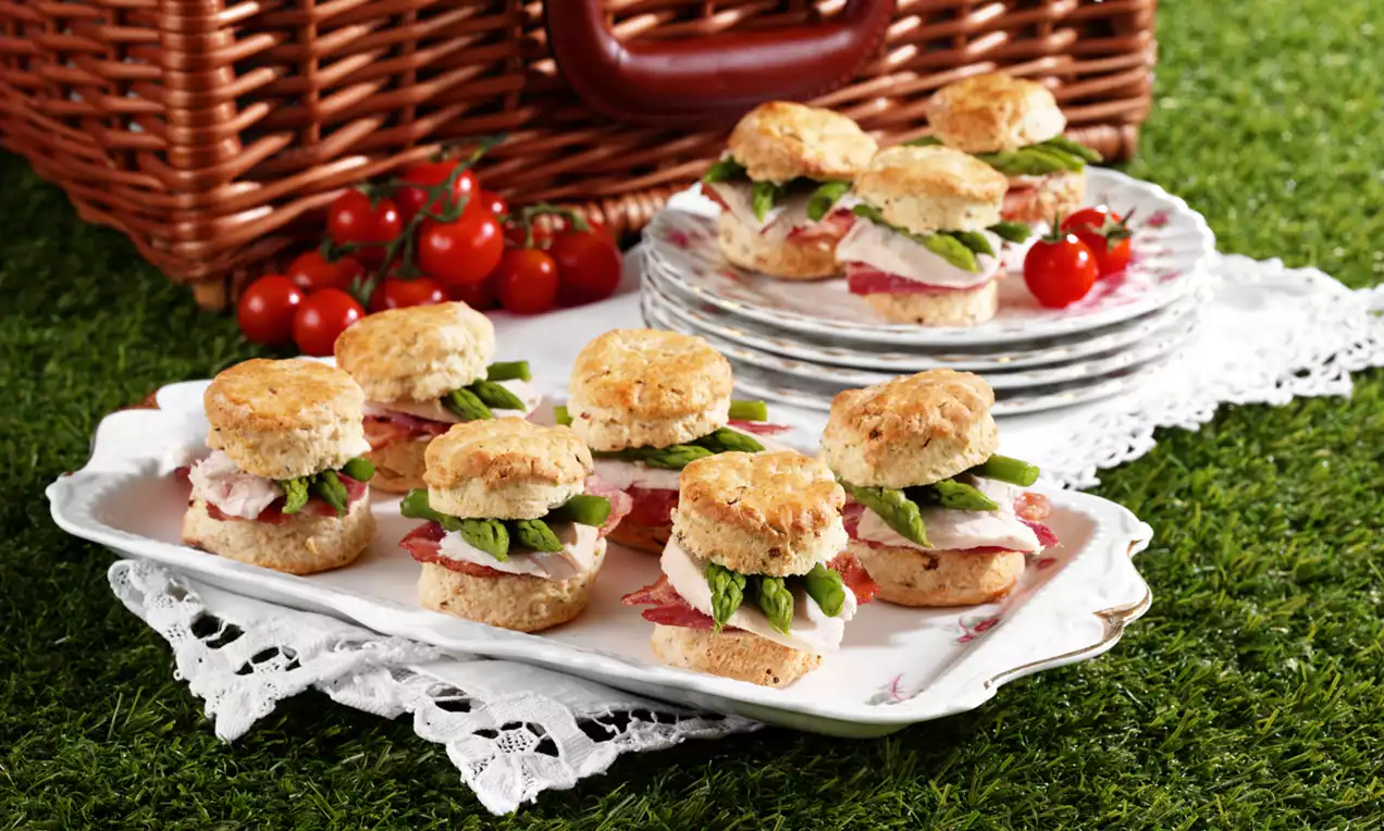 Golden scones layered with slices of ham, creamy filling, and crisp green asparagus spears on a white plate with lace detail placed on grass