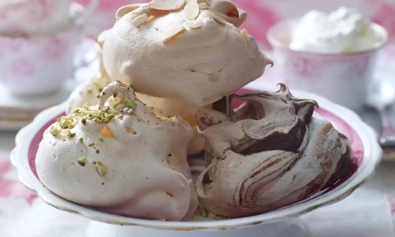Meringues aériennes aux teintes crème et marron marbrées, parsemées d’éclats de pistaches et amandes sur un plat en porcelaine blanche à bord doré
