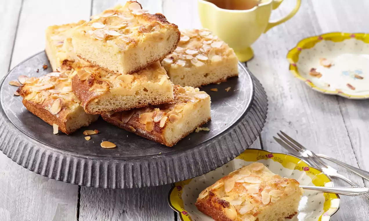 Saftige Butterkuchenstücke mit goldbrauner Kruste und gerösteten Mandelblättchen auf einer grauen Platte, umgeben von einer Teetasse und Tellern