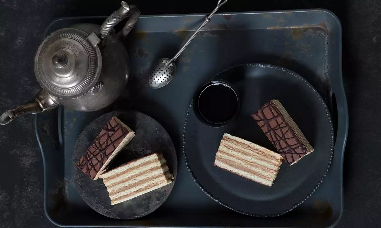 Dobošovy řezy složené z vrstev světlého piškotu a čokoládového krému, vrchní poleva s kakaovým vzorem, servírované na tmavých talířích