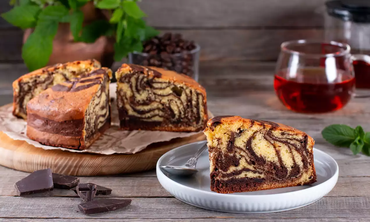 Marble cake with swirled chocolate and vanilla patterns, golden-brown crust, served on a white plate next to chocolate pieces and tea