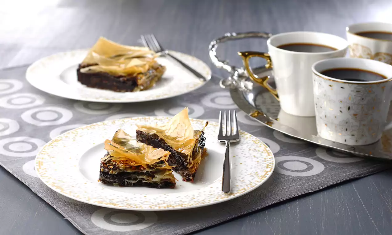 Üçgen dilimlenen supanglenin koyu, nemli dokusu, altın rengi gevrek yufka katlarıyla kontrast oluşturuyor, yanlarda kahve fincanlarıyla sunulmuş