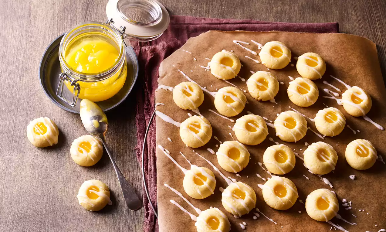 Kleine helle Plätzchen mit gelber Zitronenfüllung auf braunem Backpapier, daneben ein Glas Lemoncurd und ein Löffel auf rustikalem Holztisch