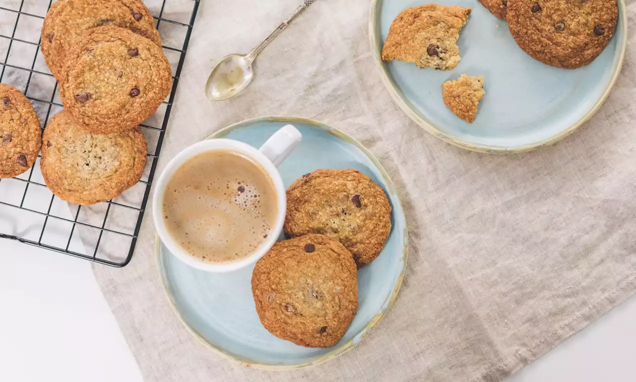 Cookies doradas con bordes crujientes y centro suave, salpicadas de chips de chocolate sobre platos celestes con taza de café espumoso
