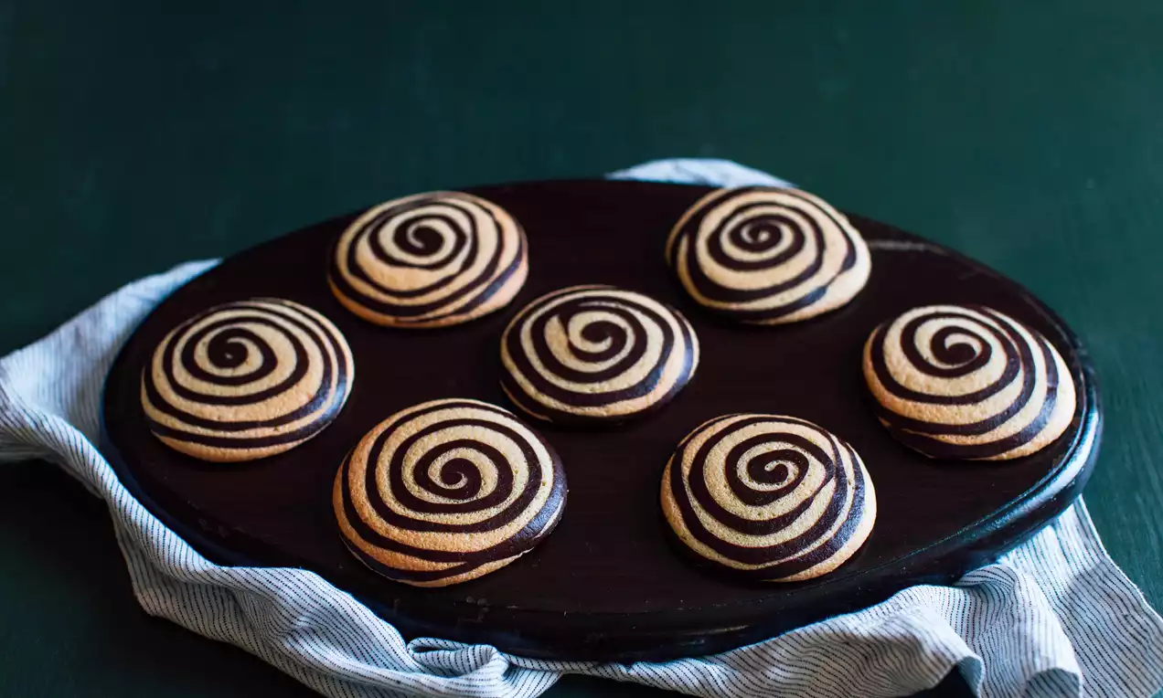 Koyu ahşap bir tabakta, kumaş üzerine yerleştirilmiş, kıvrımlı siyah ve altın rengi desenlerde yuvarlak tatlılar.