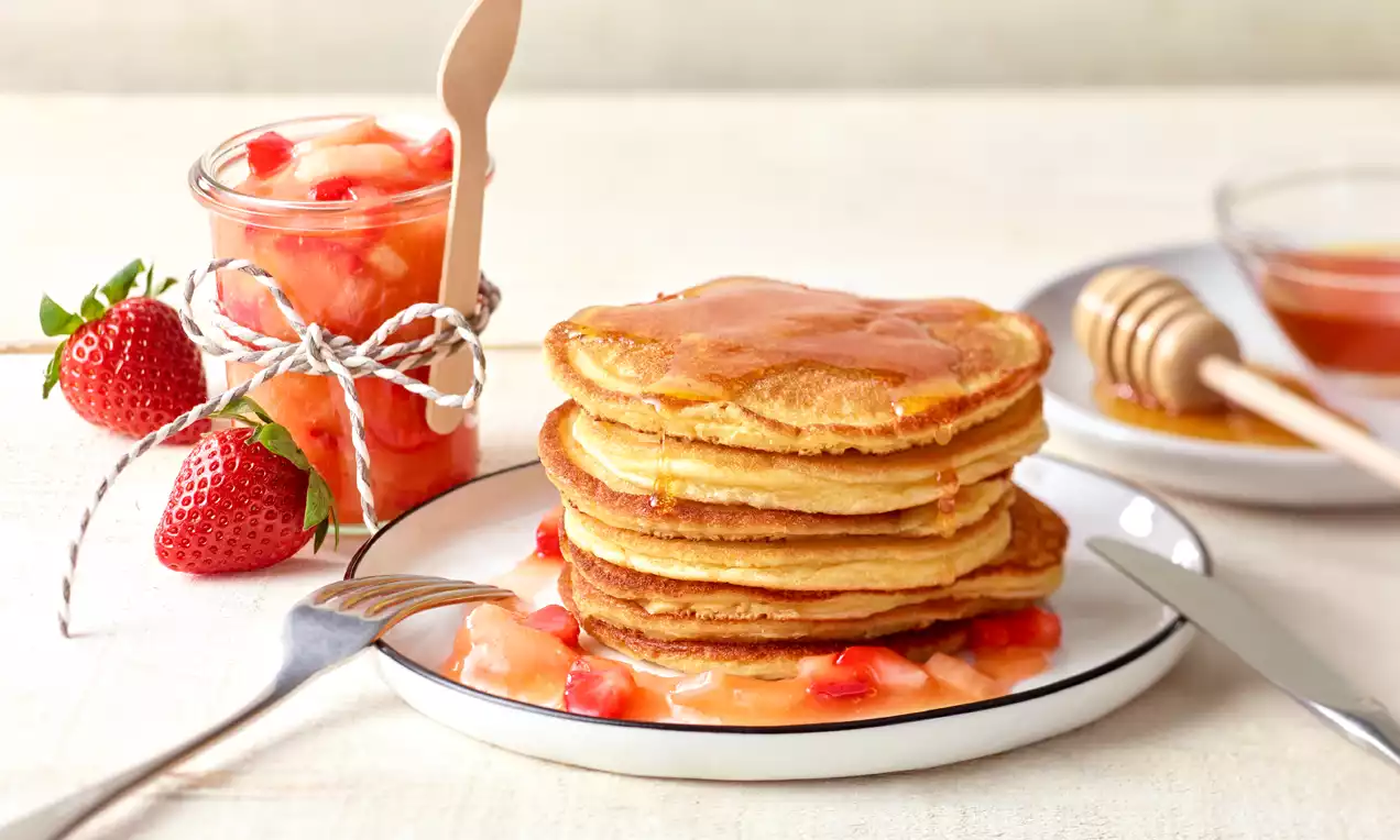 Goldbraune Pancakes mit glänzendem Erdbeer-Rhabarber-Kompott, auf einem weißen Teller neben frischen Erdbeeren und Honig