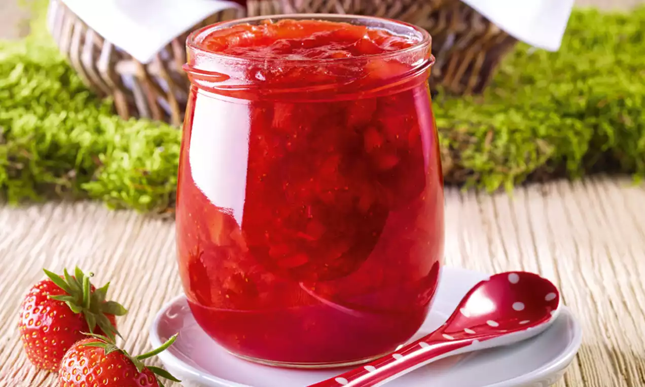 Helderrode aardbeienconfituur met glanzende en grove textuur in een glazen pot op een wit bord met een rode lepel en verse aardbeien