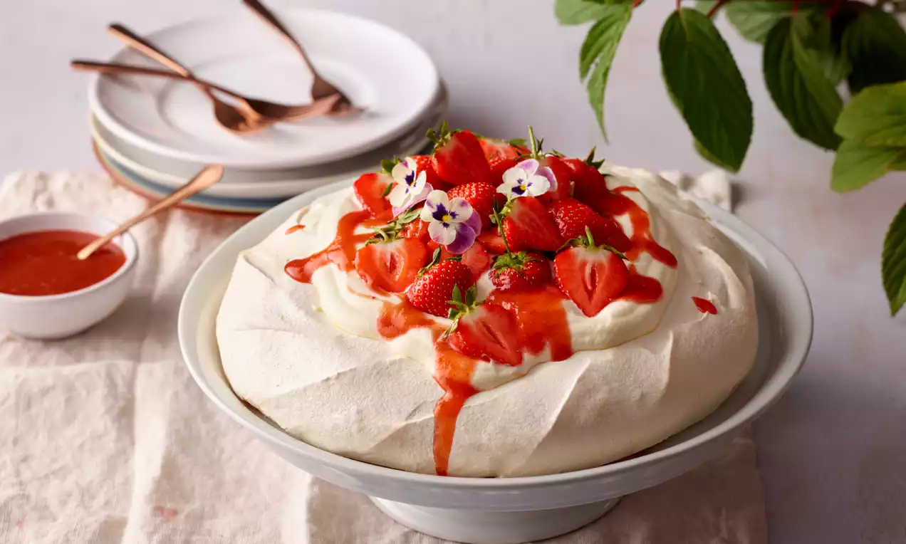 Knusprige, weiße Baiserschale gefüllt mit Sahne, frischen Erdbeeren, Rhabarbersauce und essbaren Blüten auf einem hellen Teller