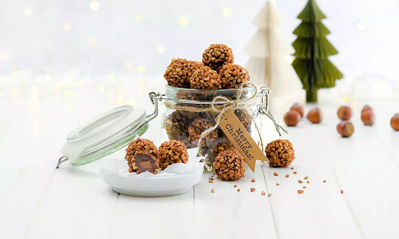 Kleine kugelige Pralinen mit braunen Nussstücken bestreut, teils mit Schokolade gefüllt, in und neben einem Glas mit Holzanhänger arrangiert