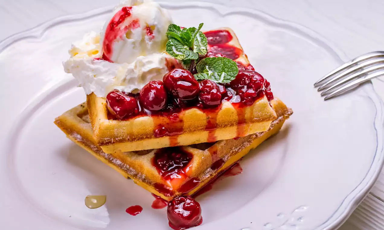 Vafe aurii stratificate cu înghețată albă, topping de sos roșu de vișine, fructe întregi lucioase și frunze de mentă pe o farfurie albă