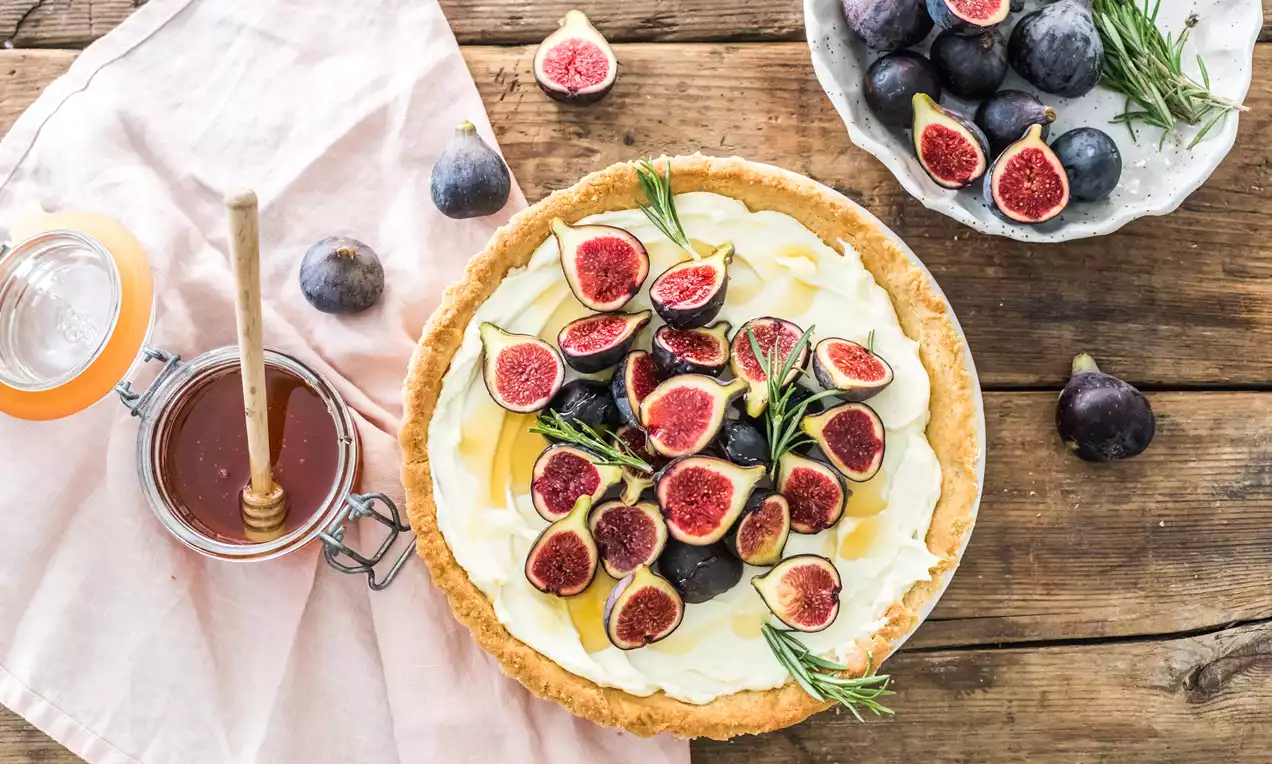 Tarta con base dorada y crema blanca cubierta de mitades de higos frescos rojo intenso y decorada con ramitas de romero sobre una mesa de madera