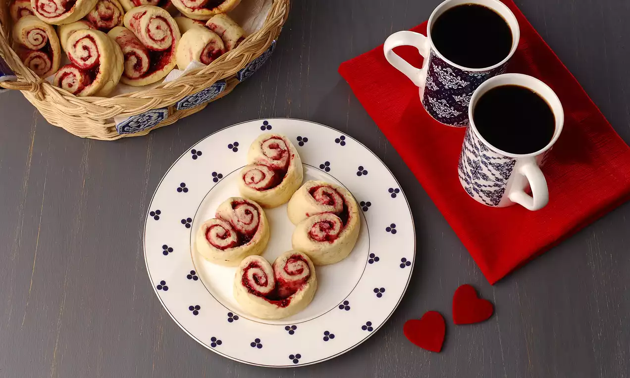 Altın rengi ince hamurla sarılmış, kalp şeklinde ahududu dolgulu kurabiyeler, desenli bir tabakta, kahve ve kırmızı dekorlarla servis edilmiş