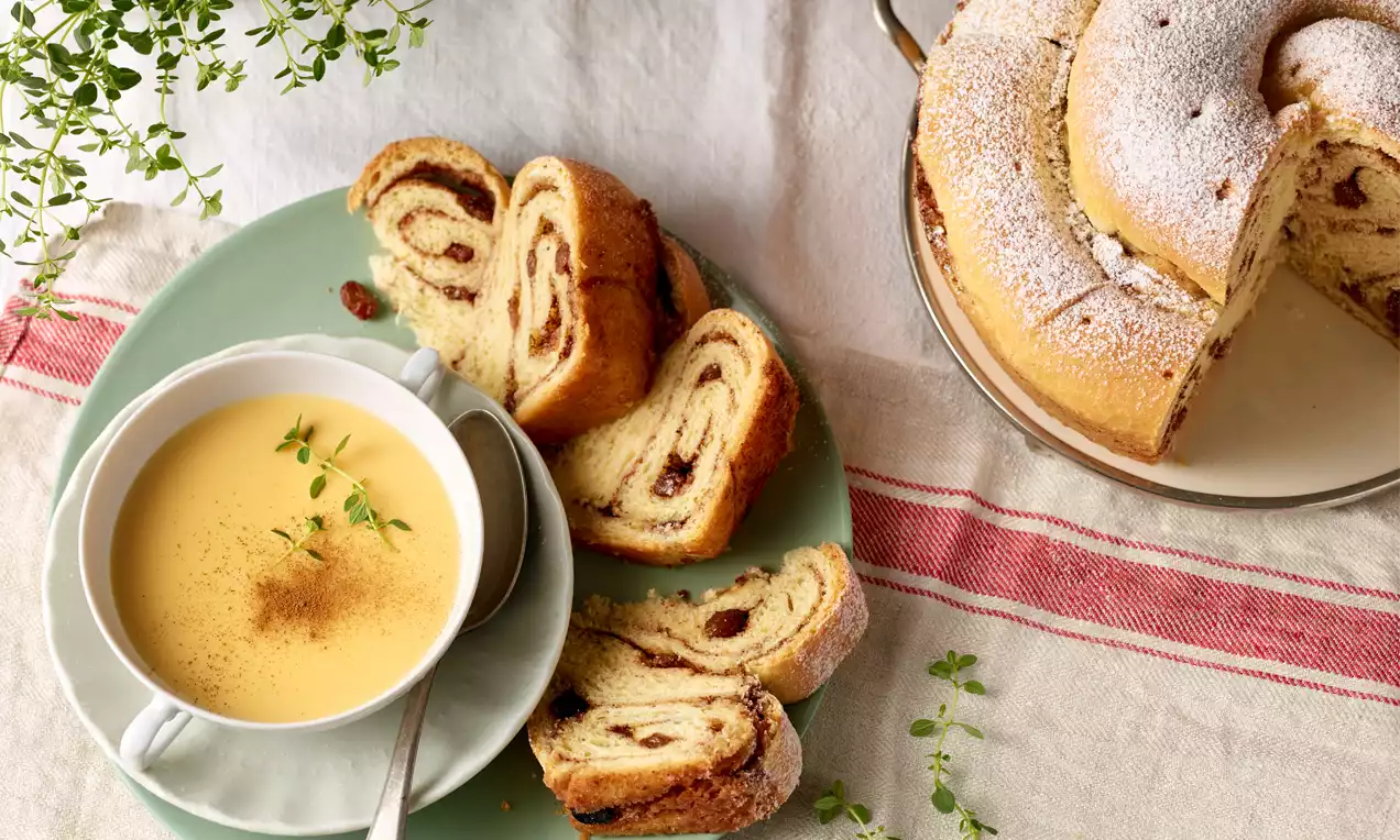Kärntner Reindling mit goldbrauner Kruste, spiralförmiger Füllung und Staubzucker, neben einer Tasse gelber Suppe mit Kräutergarnitur