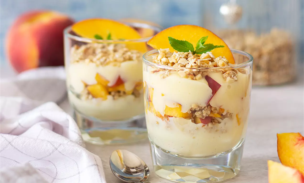 Слоест крем парфе в прозрачна чаша с бледожълт крем, сочни парченца праскова и златиста гранола, декориран с резен праскова и листенце мента
