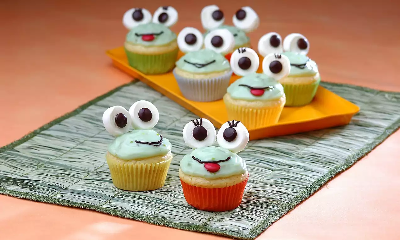 Yeşil krema kaplı kurbağa yüzü şekilli cupcakeler, şeker gözler ve detaylı süslemelerle yeşil dokulu bir örtü ve turuncu bir tabakta düzenlenmiş