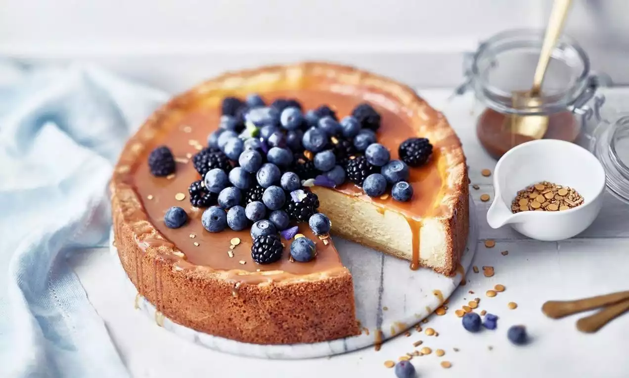 Gyldenbrun ostekake med glatt karamellglasur, toppet med friske blåbær og bjørnebær, på hvit marmortallerken med nøttepynt rundt