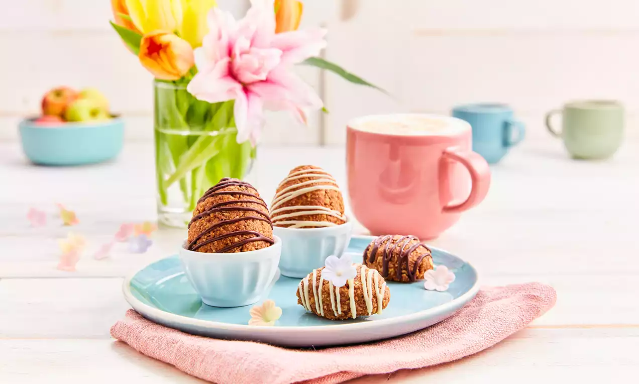 Tamnosmeđa jaja od granole s glazurom u bijelim linijama, raspoređena na plavom tanjuru s cvjetnim dekoracijama i šalicom kave