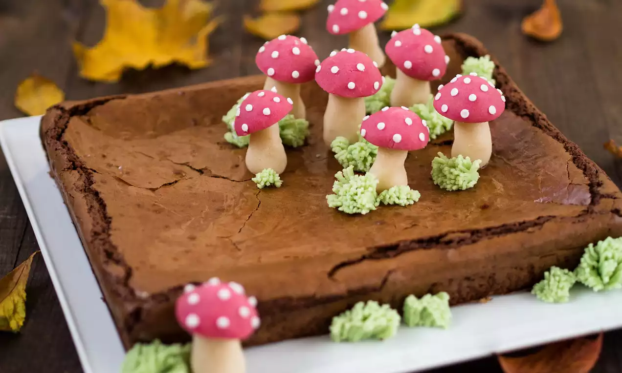 Ciemnobrązowe brownie z błyszczącą powierzchnią, udekorowane czerwonymi muchomorkami z białymi kropkami i kremowymi choinkami na białym talerzu