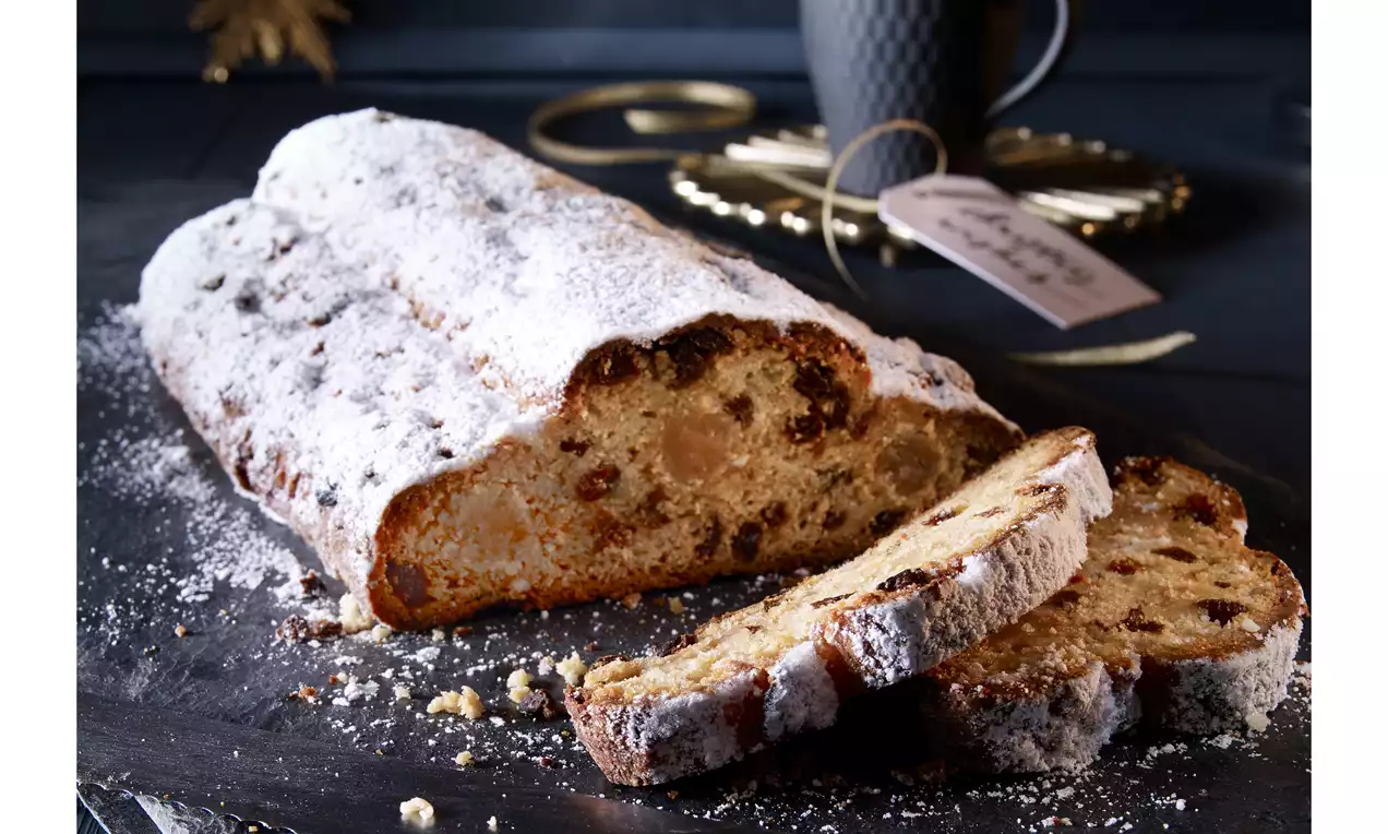 Hófehér porcukorral hintett, aranyszínű mazsolás kalács omlós szeletekkel, sötét tálcán, aranyló díszek és fekete bögre kíséretében