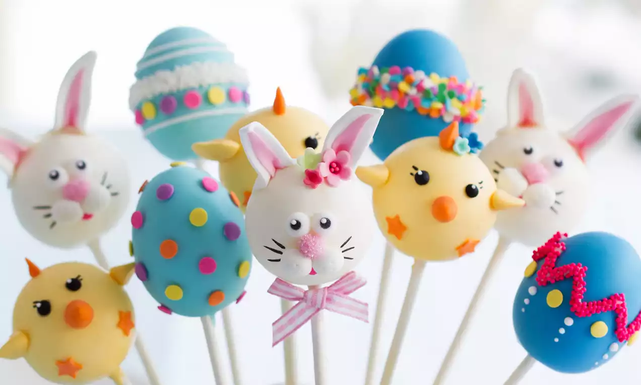 Popcakes in paasstijl op witte stokjes, versierd als konijnen, kuikens en felgekleurde eieren, geplaatst in een lichtblauw emmertje met gele linten