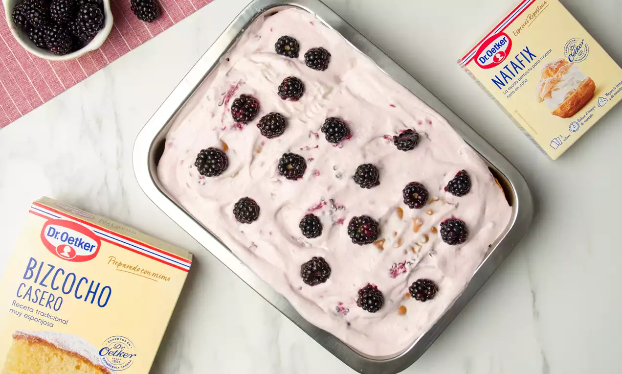 Short cake rectangular con crema suave rosa claro, trozos de bizcocho dorado y moras negras distribuidas sobre una bandeja metálica