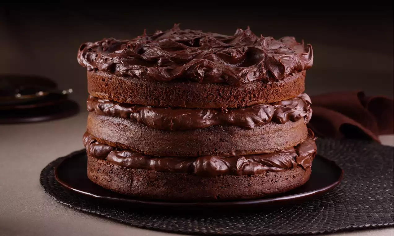 Three-layer chocolate cake with rich, glossy dark chocolate cream between each layer and swirled on top, served on a dark plate