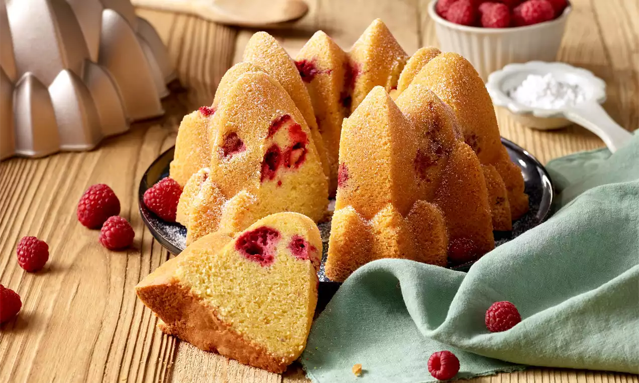 Malinová bábovka zlatavé barvy s šťavnatými malinami uvnitř, na černém talíři zdobeném čerstvými malinami a zelenou látkou