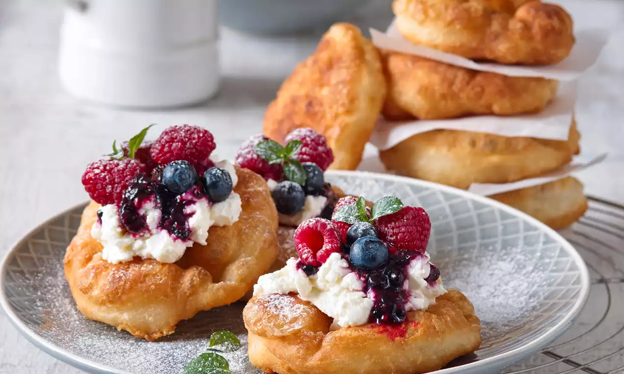 Zlatavé dozlatova smažené langoše nepravidelného tvaru s kopečky bílé ricotty, čerstvého lesního ovoce a snítkami máty na jemně zdobeném talíři